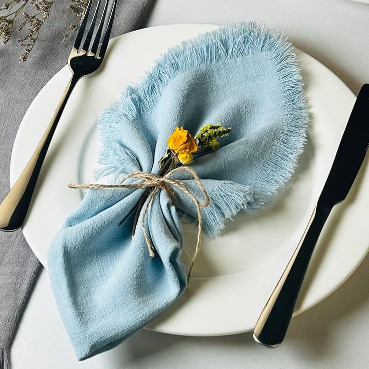 a white plate topped with a blue napkin and yellow flower next to a fork and knife