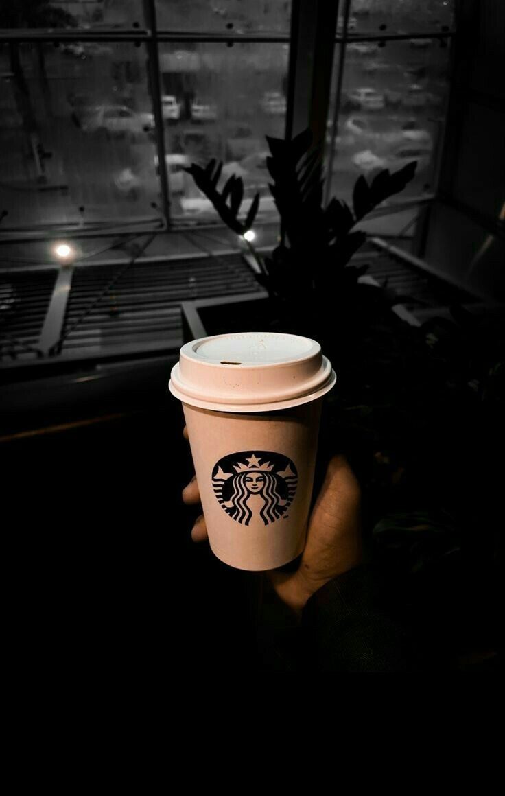 a hand holding a starbucks cup in front of a window with the city lights behind it