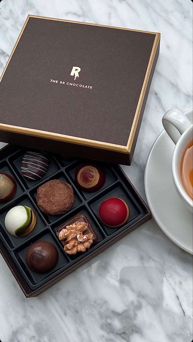 an assortment of chocolates in a box next to a cup of tea and saucer