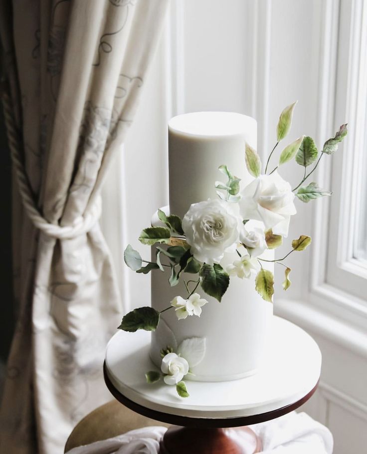 a white wedding cake with flowers on top sitting on a table in front of a window