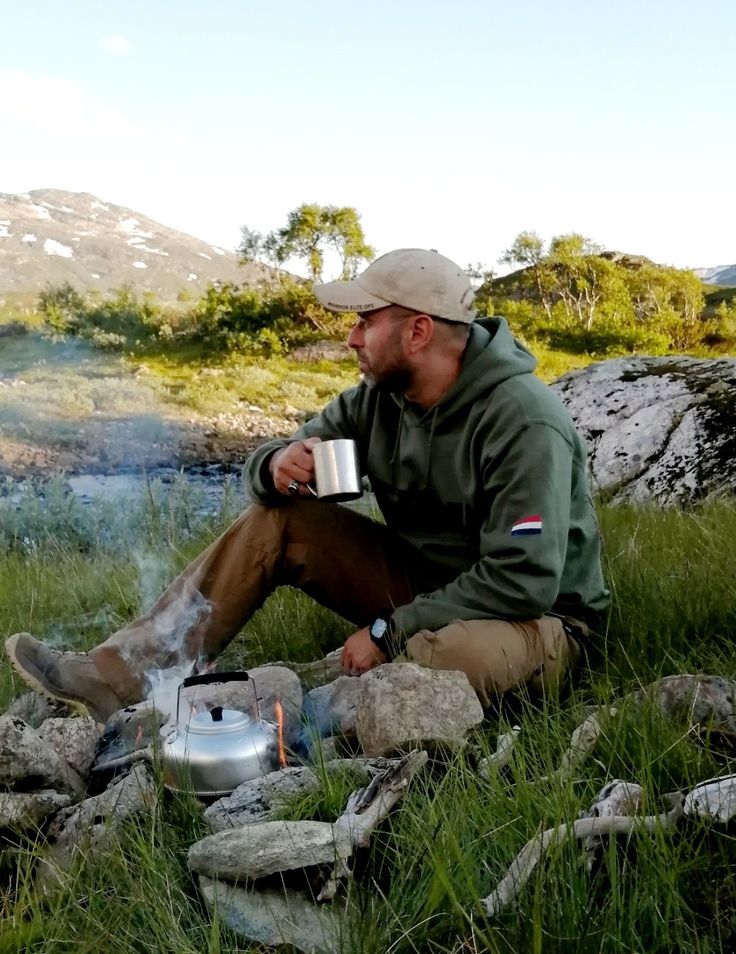 a man sitting in the grass next to a campfire with a cup on it