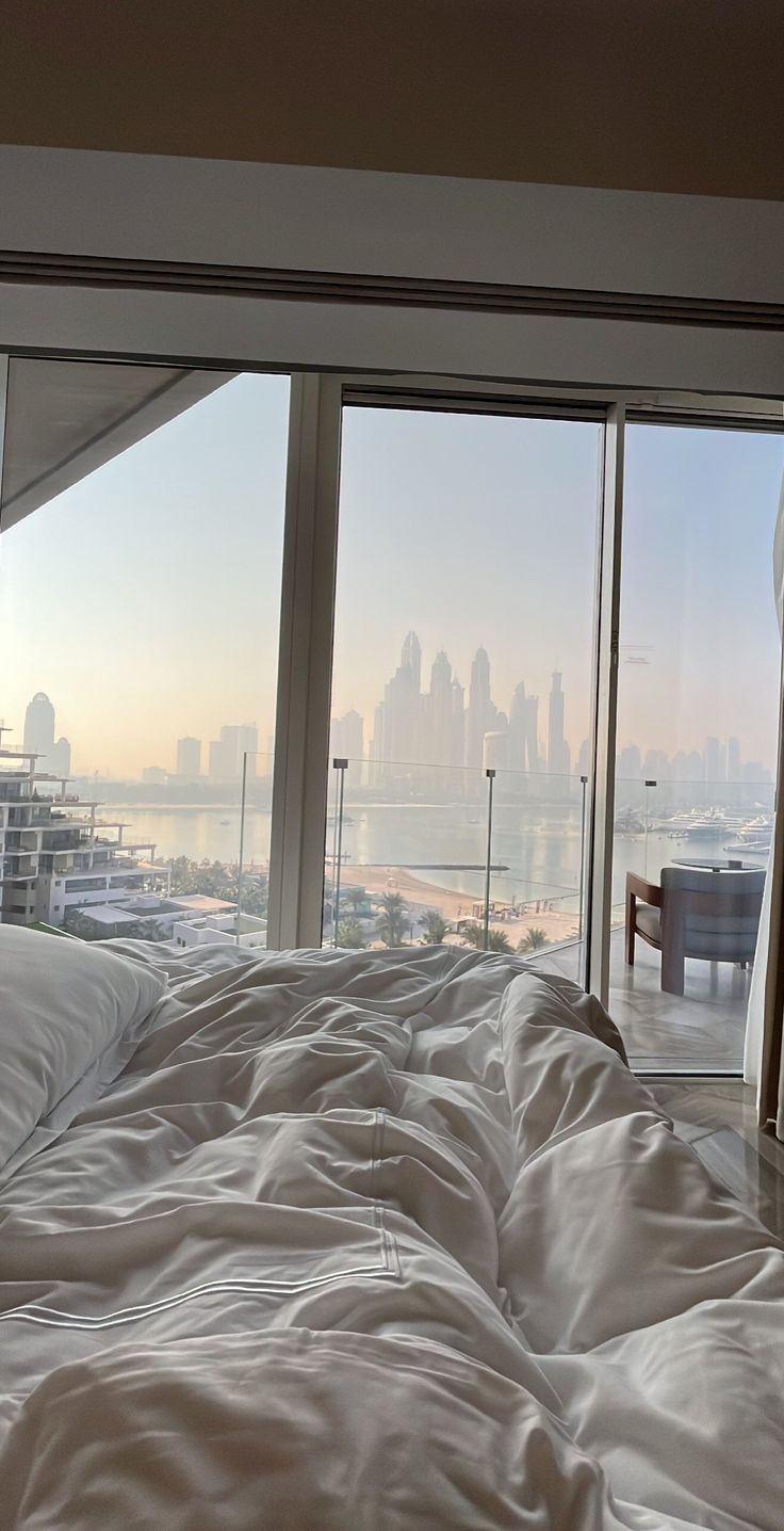 an unmade bed in front of large windows overlooking the cityscape and water