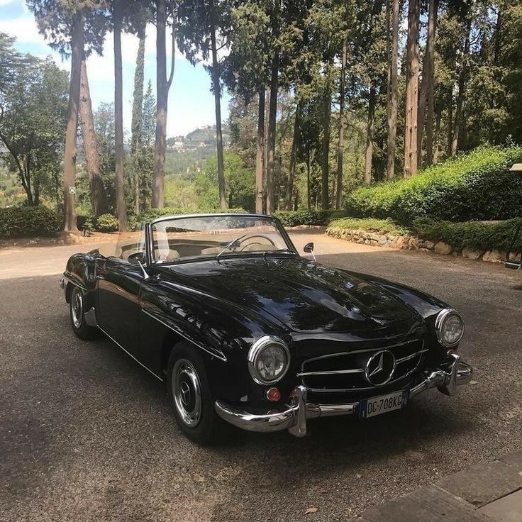 an old black car parked in a parking lot next to some trees and shrubbery