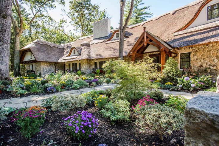 a stone house with thatched roofs and flowers in the foreground, surrounded by trees