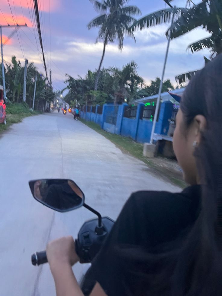 a woman riding on the back of a motorcycle down a street next to palm trees