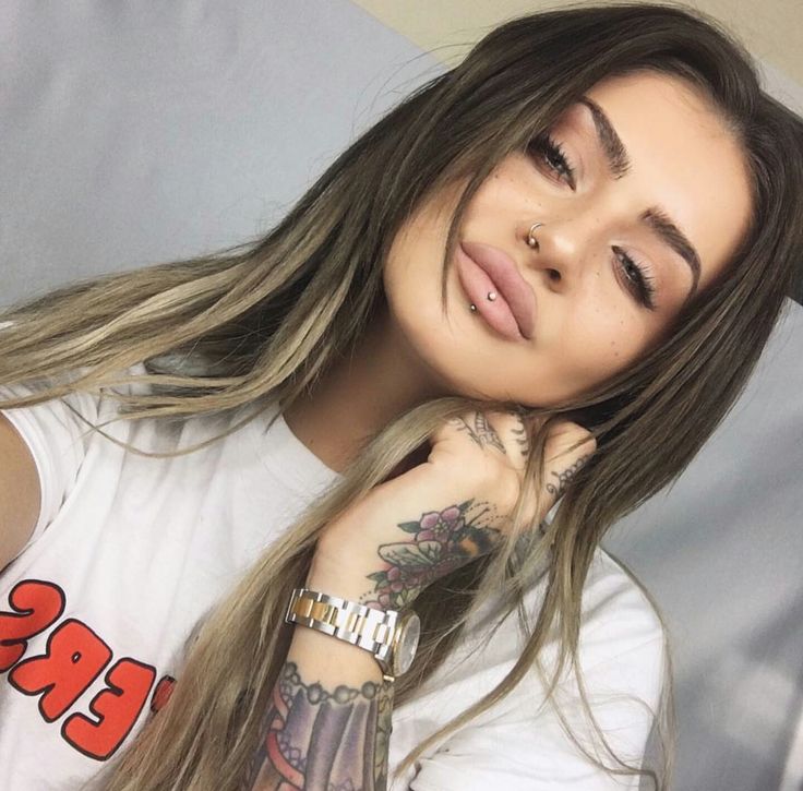 a woman with long hair and tattoos on her arm, wearing a white t - shirt that says 3 piercings for $ 100