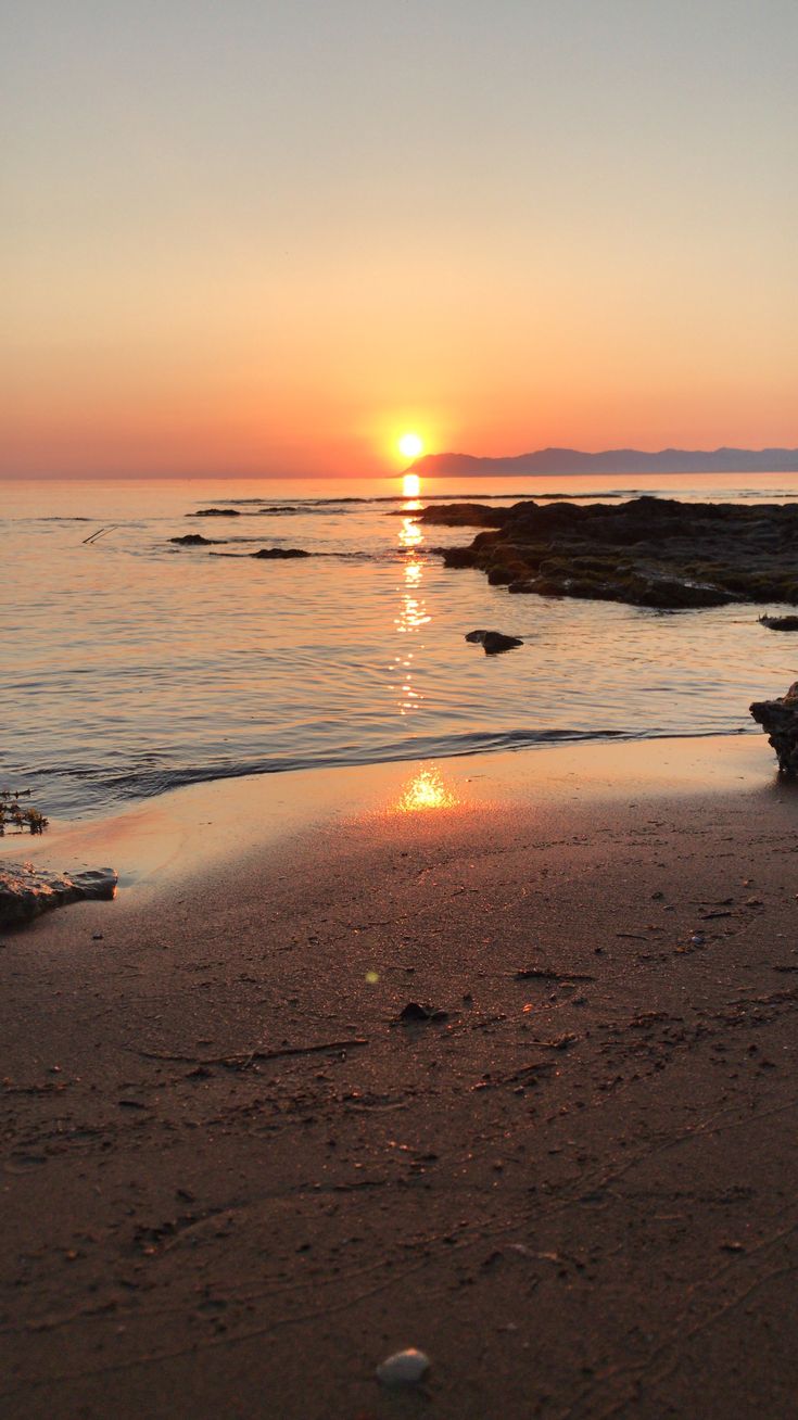 the sun is setting over the water at the beach with footprints on the sand and rocks