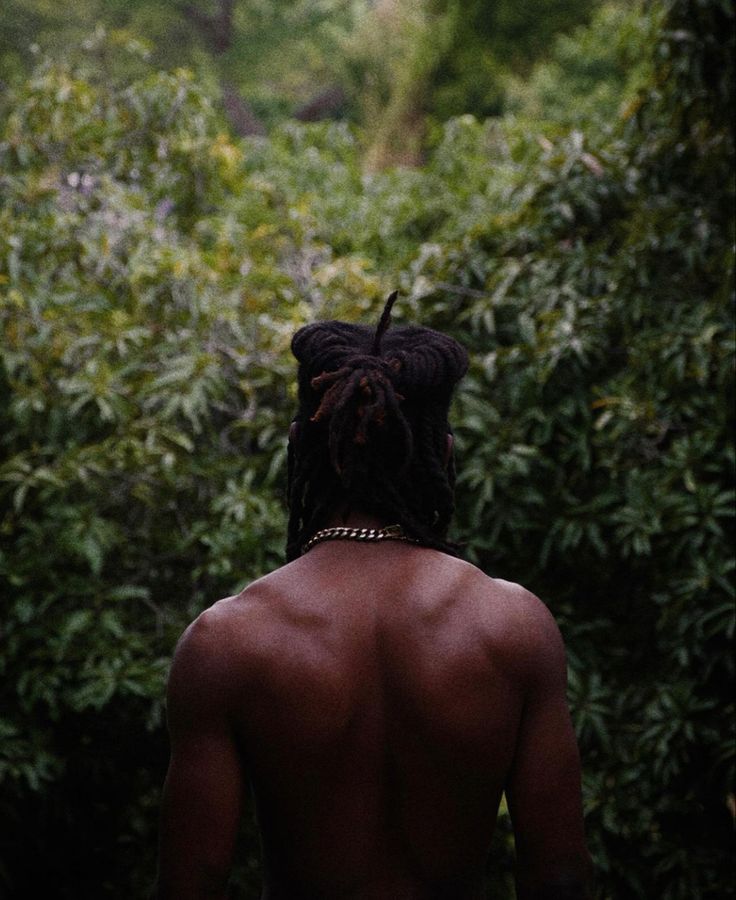 a man with dreadlocks standing in front of trees and bushes looking at something