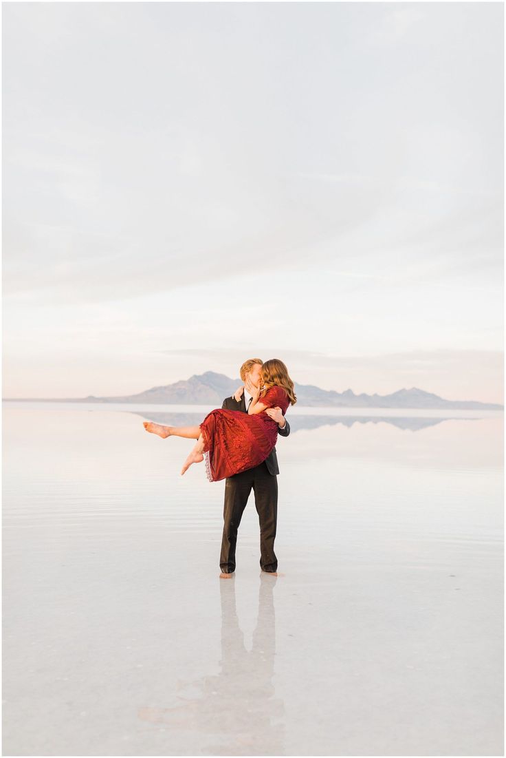 a man holding a woman in his arms while standing on the water's edge