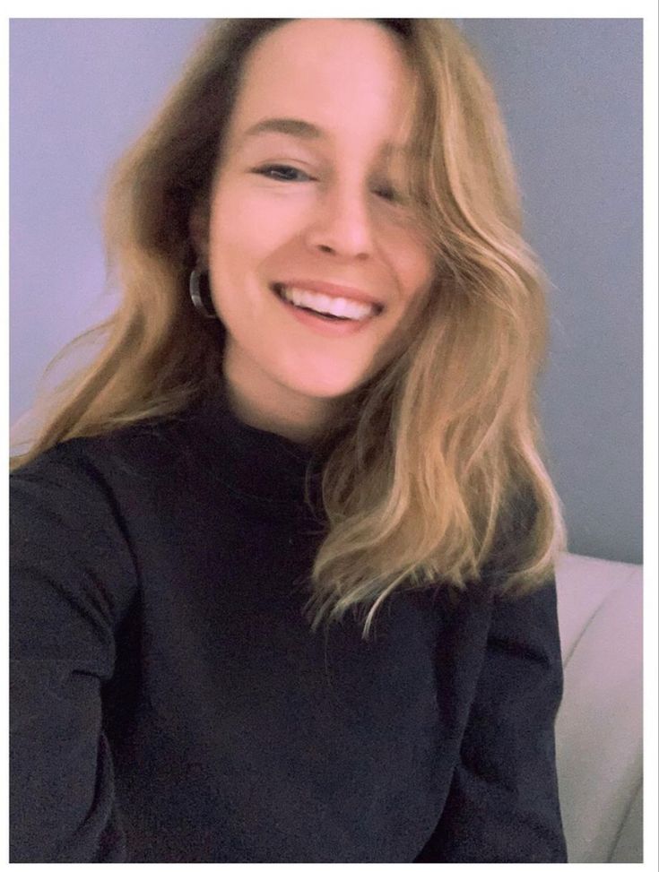 a woman with blonde hair smiling and wearing a black turtle neck sweater, sitting on a white couch