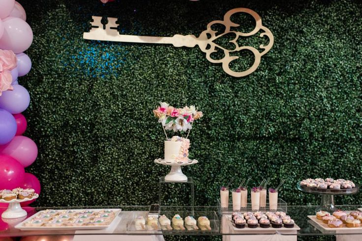 a dessert table with cupcakes, cake and flowers on it in front of a green wall