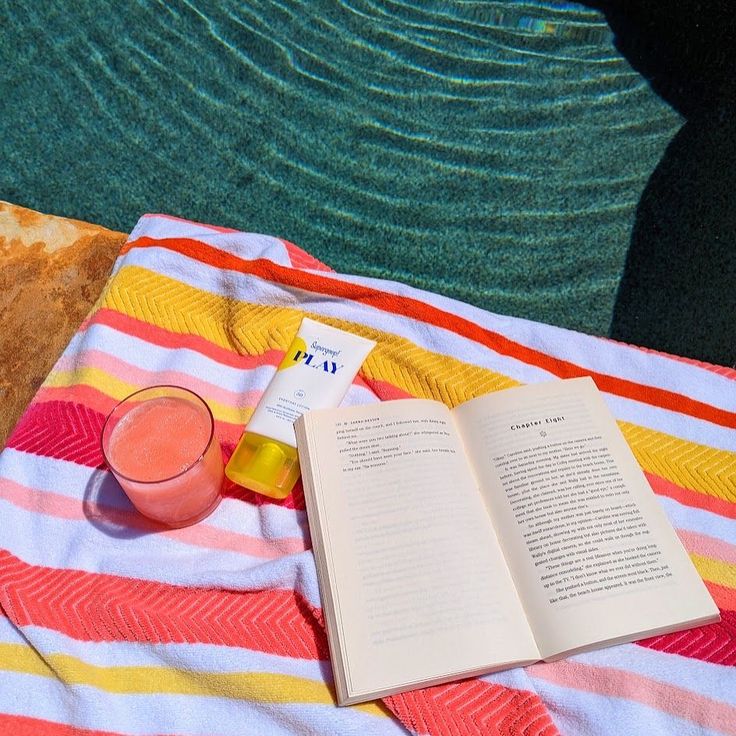 an open book sitting on top of a beach towel next to a glass of juice