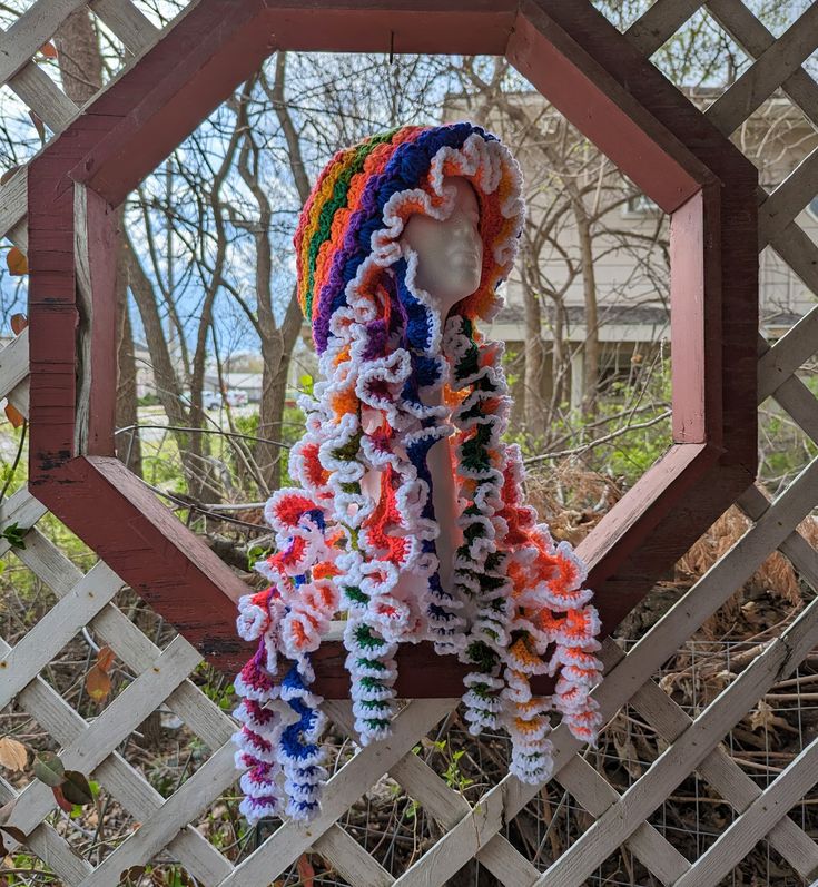 a multicolored crocheted scarf hanging on a wooden fence with trees in the background