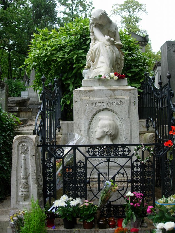 the statue is surrounded by many flowers and greenery in front of an iron fence