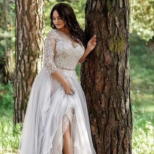 a woman standing next to a tree wearing a dress with long sleeves and high slit