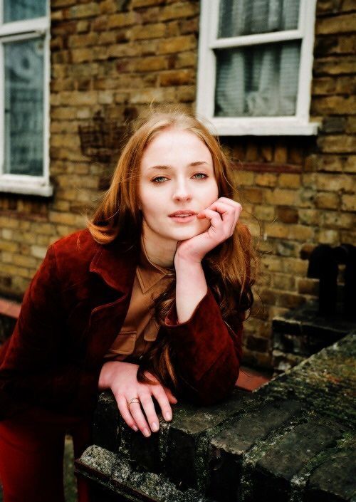 a woman sitting on a bench in front of a brick building with her hand under her chin