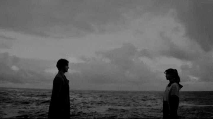 two people are standing on the beach looking out at the ocean under a cloudy sky