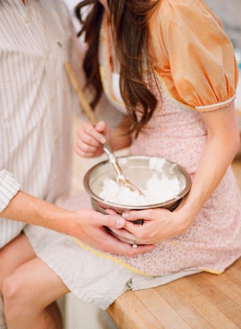 a man and woman sitting on a kitchen counter holding a bowl with food in it