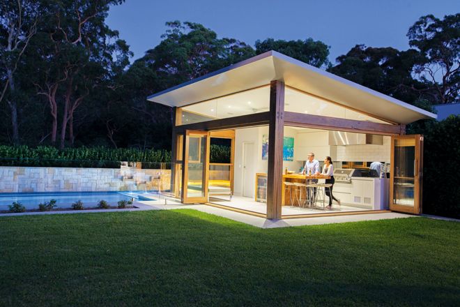 a man standing in the back yard of a house at night with an outdoor kitchen and dining area