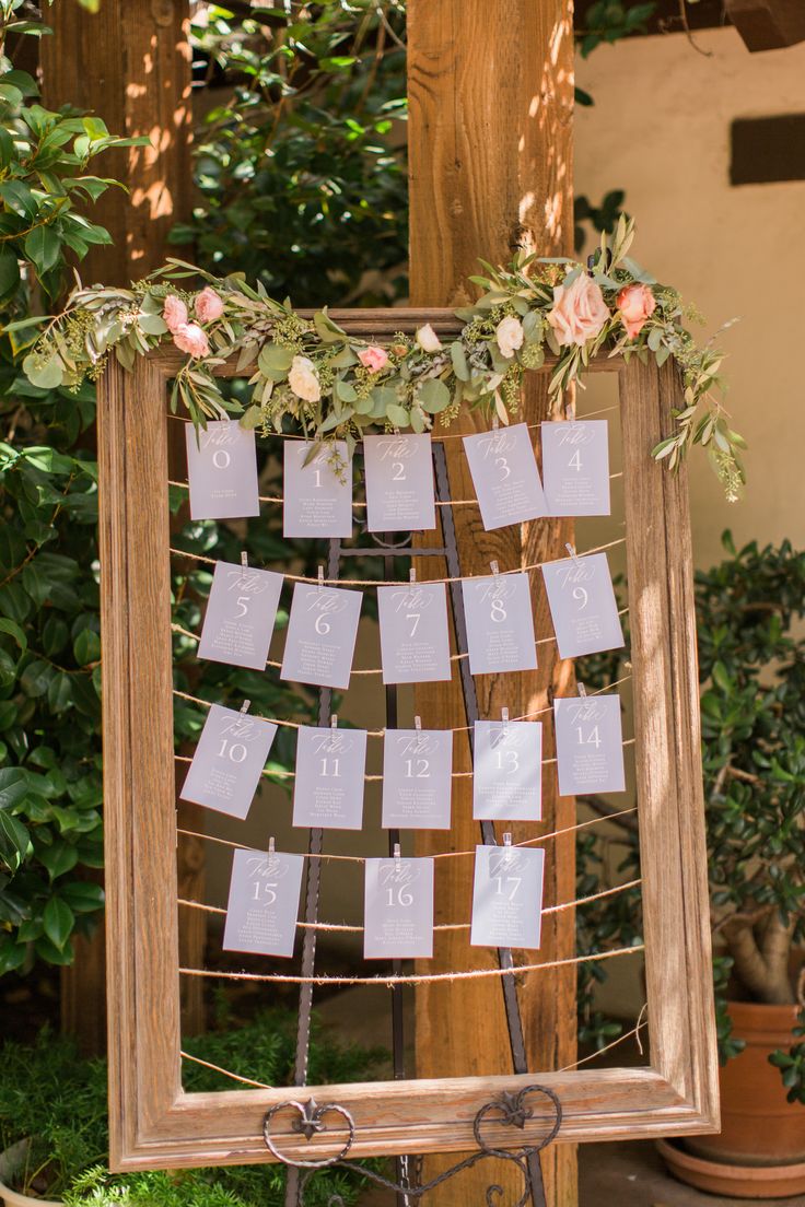 a wooden frame with seating cards attached to it and greenery around the edges is decorated with flowers