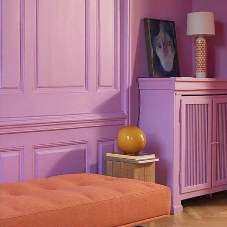 an orange ottoman in front of a purple wall with a portrait on it and a lamp next to it