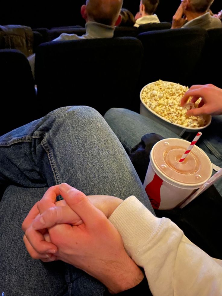 two people sitting on a couch with popcorn and drinks in their hands, watching movies