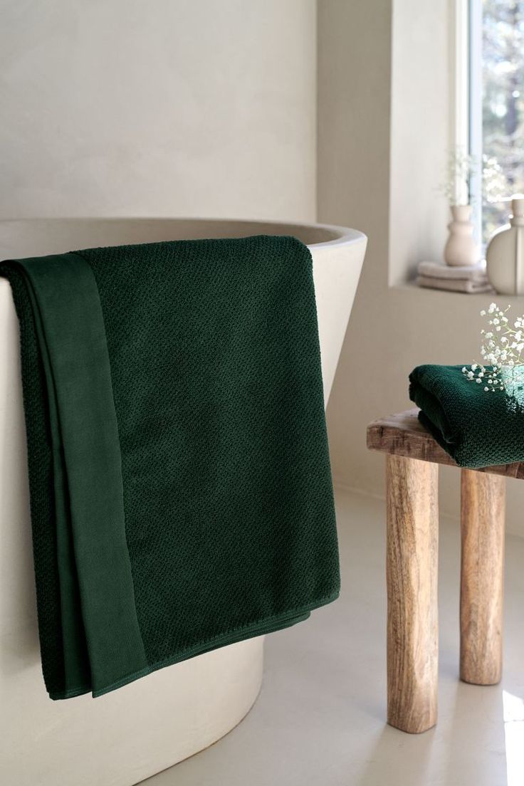 a green towel is sitting next to a white bathtub and wooden stool in the bathroom