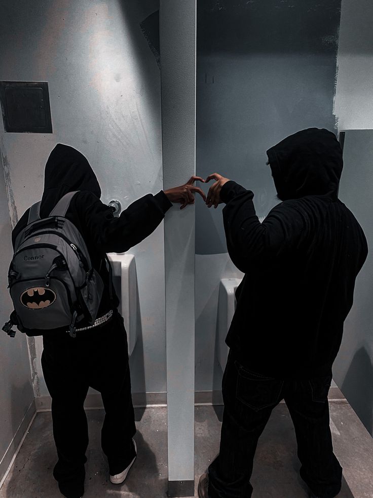 two people in black hoodies are standing near a toilet and looking at each other