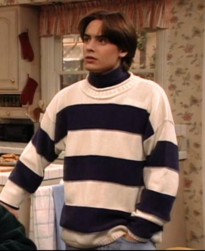 the young man is standing in his kitchen wearing a striped sweater and jeans, looking off into the distance