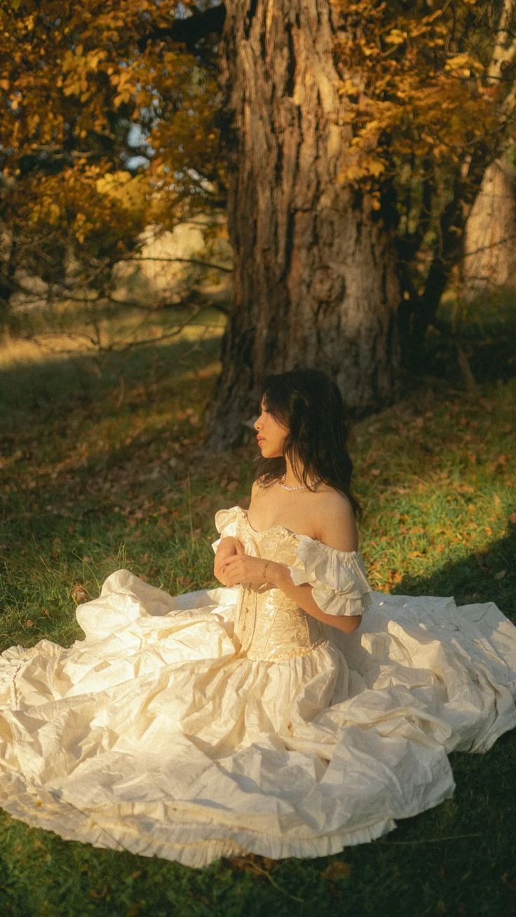 a woman in a white dress sitting on the ground next to a tree and holding a bow