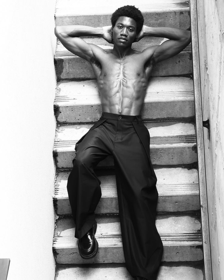 a man with no shirt is sitting on some stairs and posing for the camera while holding his hands behind his head