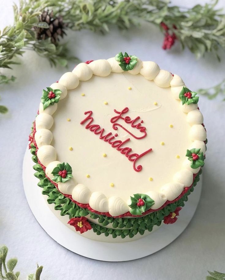 a white cake decorated with holly leaves and the words happy holidays written on it, surrounded by greenery