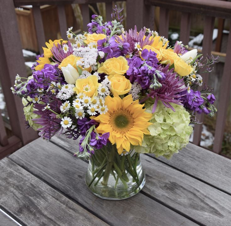 a glass vase filled with yellow and purple flowers on top of a wooden table next to snow covered ground