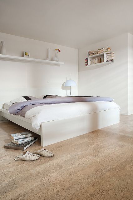 a bedroom with white walls and flooring has a large bed, bookshelf, and lamp