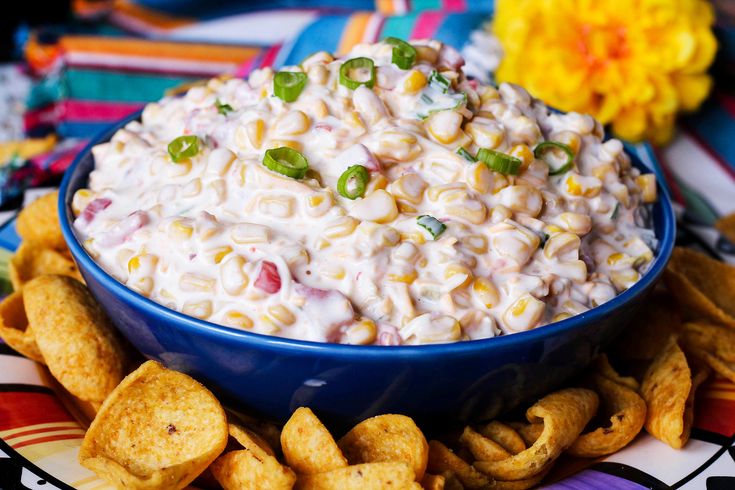 a blue bowl filled with corn salad surrounded by chips on a colorful table cloth and flowers