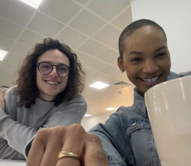 two women sitting at a table with coffee cups in front of them, one smiling and the other pointing to the camera