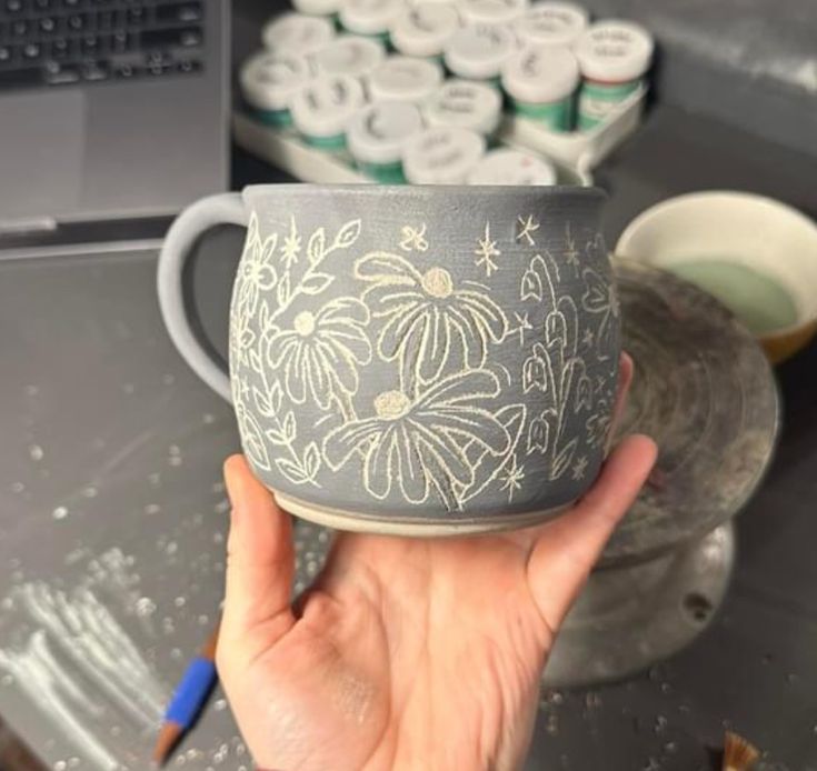 a hand holding a cup with flowers painted on it next to other cups and saucers