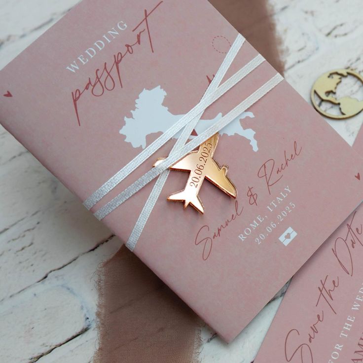 two pink wedding cards with gold airplane brooches on them, sitting next to each other