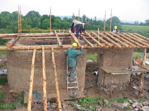 two men working on the roof of a house under construction with wooden beams and straw bales