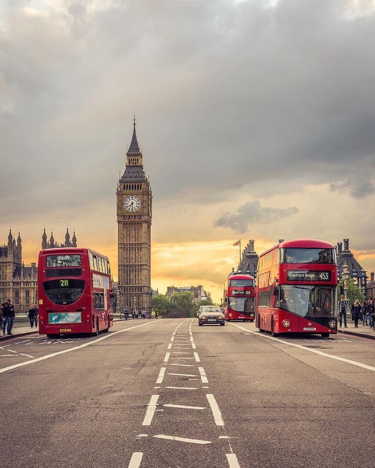 two red double decker buses driving down the street in front of big ben and the palace of westminster