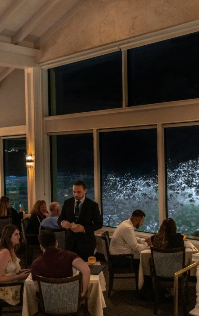 a group of people sitting at tables in a restaurant with large windows looking out on the water