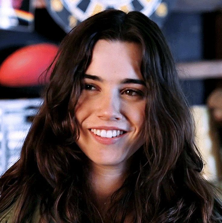 a close up of a person wearing a jacket and smiling at the camera with a clock in the background