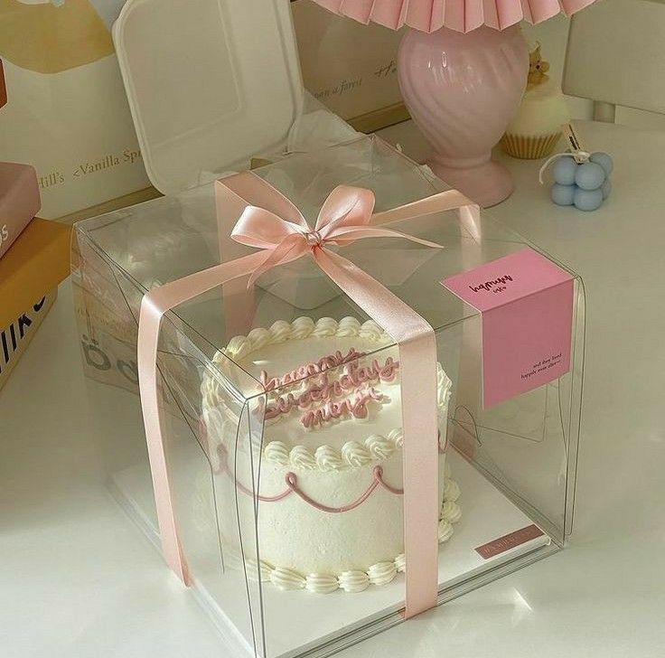 a pink and white cake in a clear box on a table next to a lamp