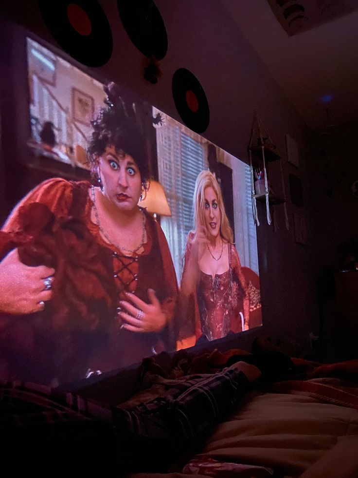 a person laying on a bed in front of a large screen with two women standing next to each other