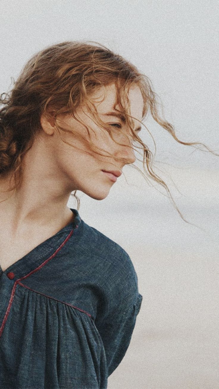 a woman standing on the beach with her hair blowing in the wind and looking off to the side