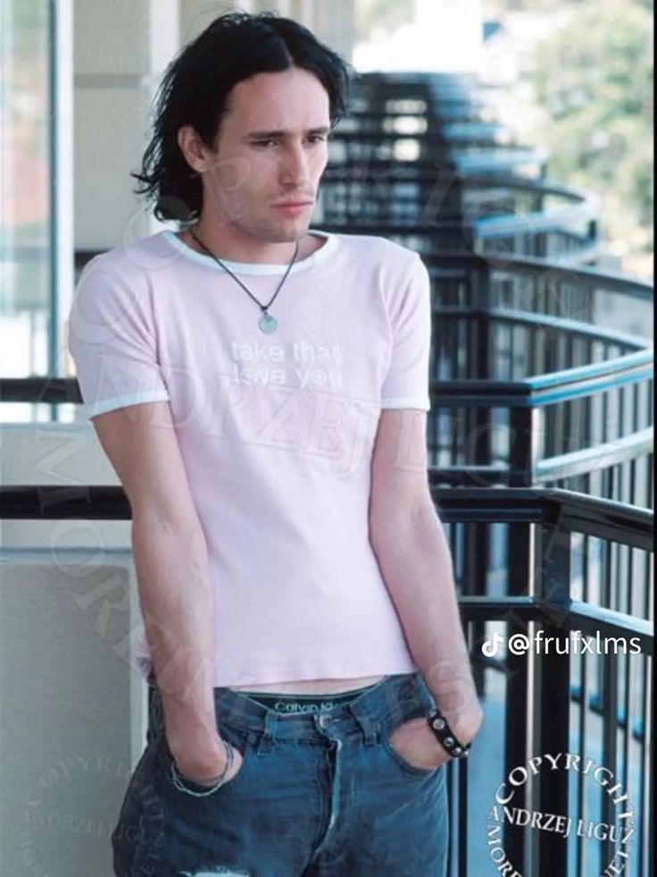 a man with long hair wearing a t - shirt and jeans standing next to a railing