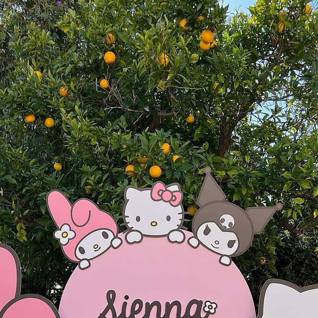 a sign with hello kitty on it in front of an orange tree and some trees