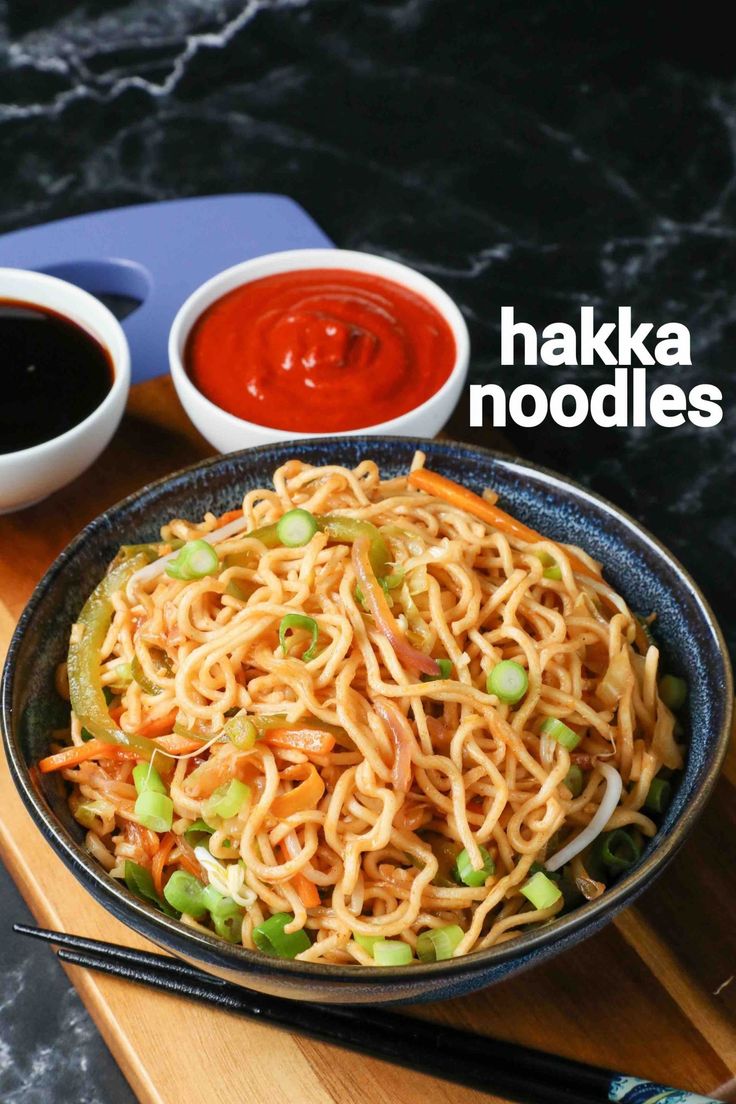 a bowl filled with noodles and vegetables next to dipping sauces on a cutting board