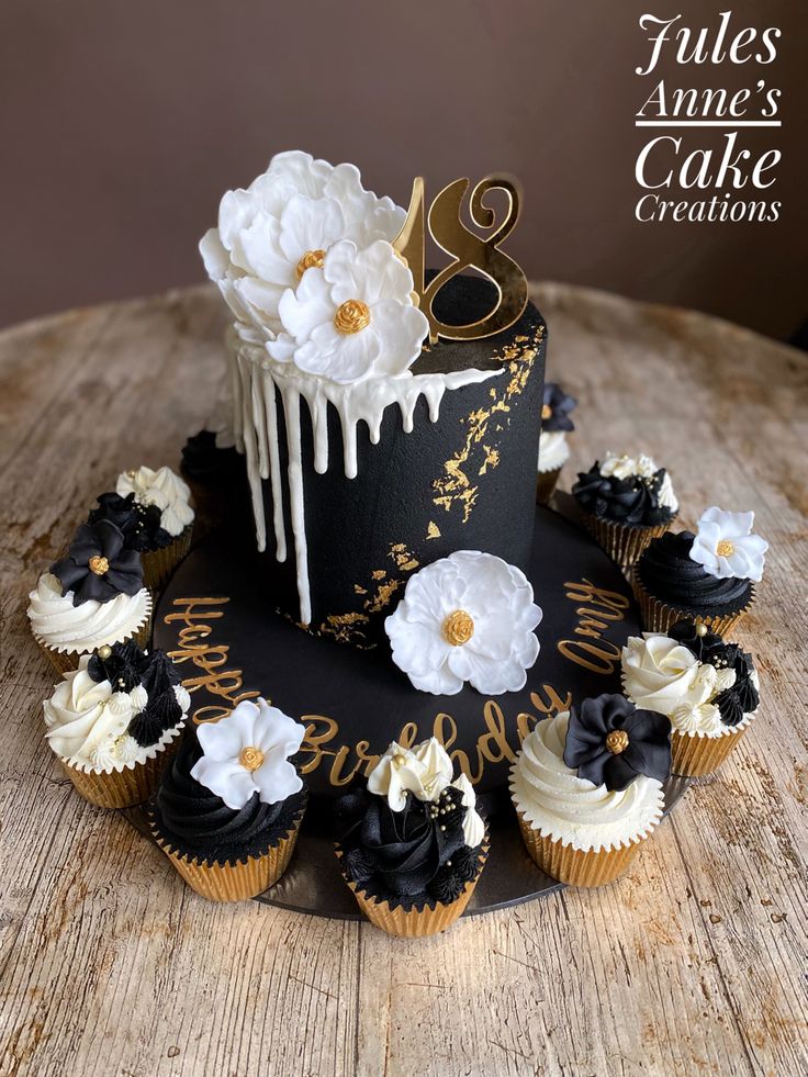 there is a black and white cake with flowers on the top, surrounded by cupcakes