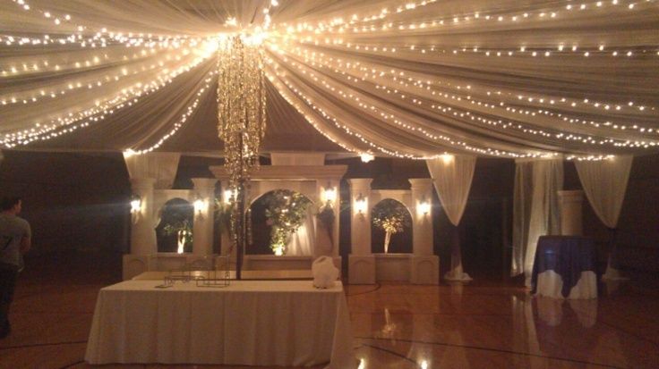 an indoor wedding venue with white drapes and lights on the ceiling, decorated with candles